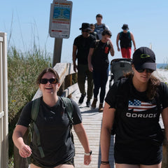 A group with backpacks, one featuring the Subtle Spearhead Tee - Tri-Blend, walks along a wooden path toward the beach on a sunny day, embodying Special Forces camaraderie.