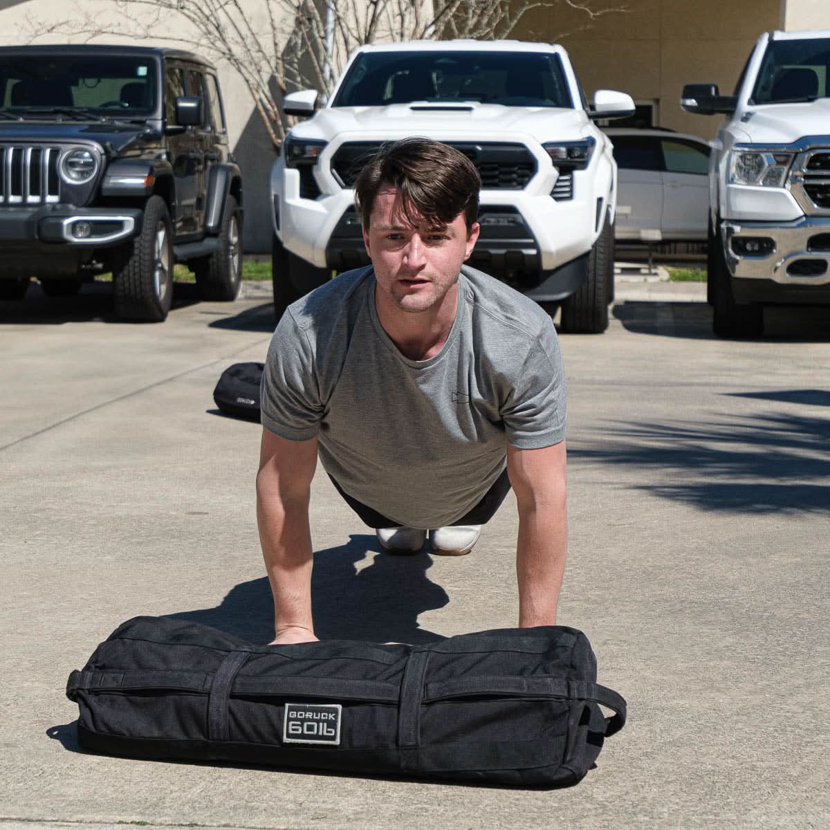 A man does push-ups outdoors wearing the Subtle Spearhead Tee - Tri-Blend, featuring the GORUCK spearhead logo, with a black sandbag on the ground and cars parked behind him.