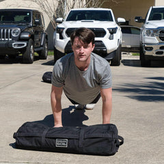Wearing the Subtle Spearhead Tee - Unisex Tri-Blend in gray, a man does push-ups on pavement near parked cars, with a GORUCK spearhead weighted bag beside him.