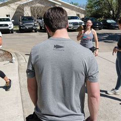 A man wearing the Subtle Spearhead Tee - Tri-Blend with the GORUCK spearhead on back is seen outdoors in a parking lot, facing away as people laugh and talk.