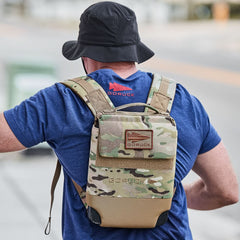 A person wearing a camo backpack and a blue Subtle Spearhead Tee - Tri-Blend walks outdoors, facing away from the camera.