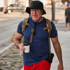 An older man wearing the Subtle Spearhead Tee - Tri-Blend in blue and red shorts strolls the sidewalk with a hat, backpack, and coffee cup—embodying everyday adventure with a Special Forces spirit.