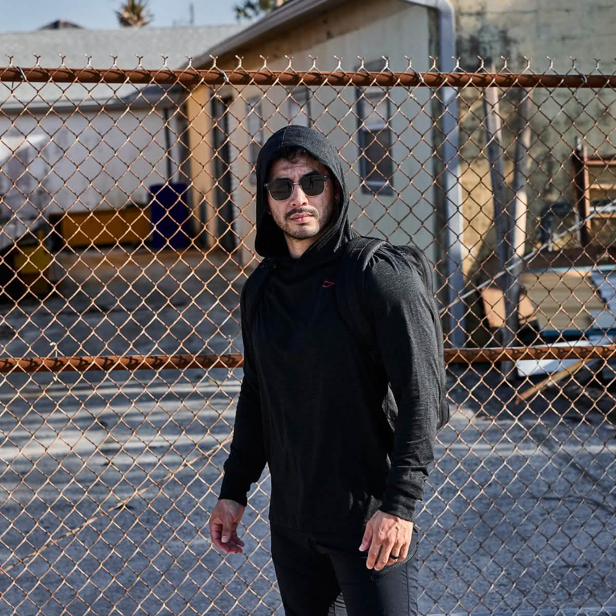 A man in a GORUCK Subtle Spearhead Hoodie - Tri-Blend and sunglasses stands near a chain-link fence against an urban backdrop.