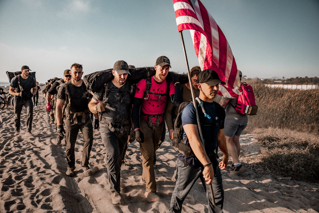 Group of people hiking on a sandy path, carrying heavy gear and an American flag, under a clear sky.