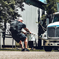 Muscular man wearing GORUCK rucksack pulling a truck with resistance ropes outside a metal building