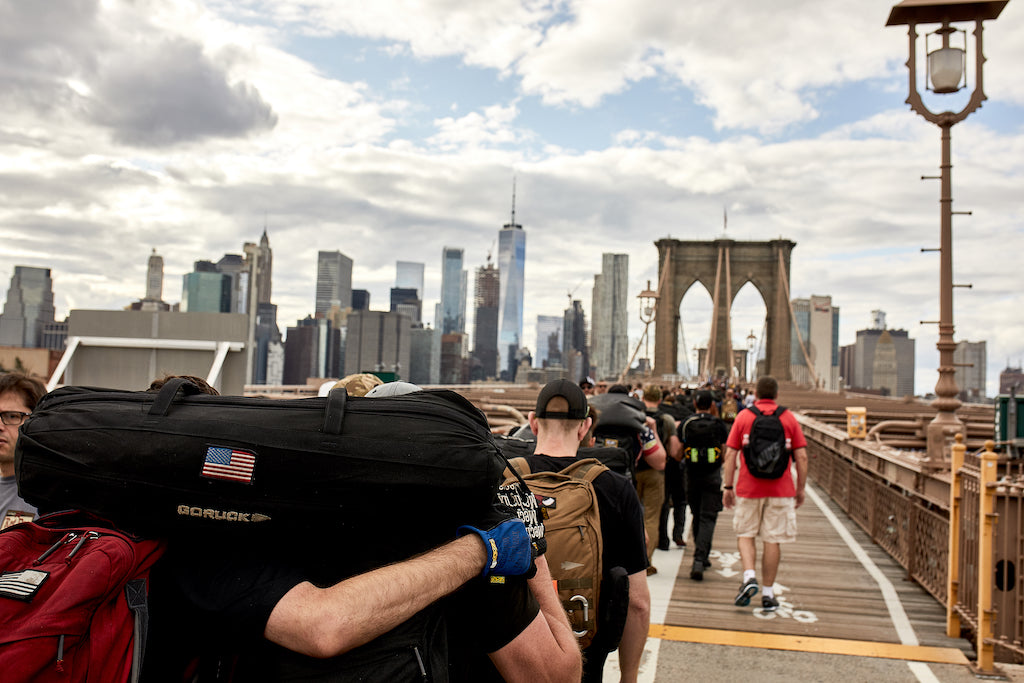 A group of people carrying backpacks walk across the Brooklyn Bridge toward the Manhattan skyline.