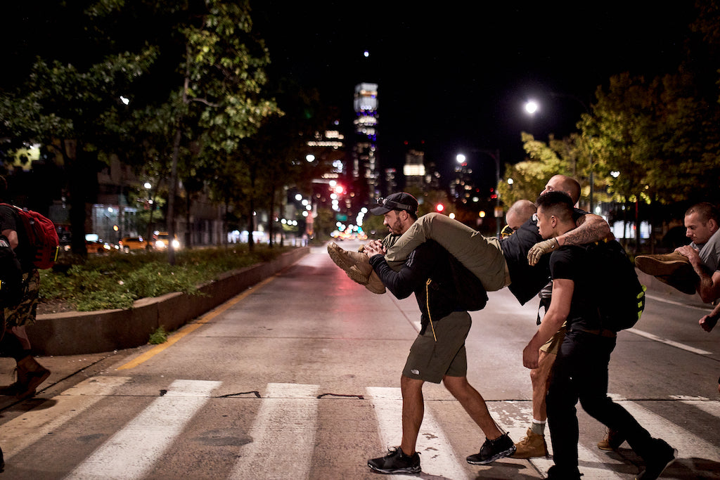 Four men cross a city street at night, two carrying others on their backs, under streetlights.