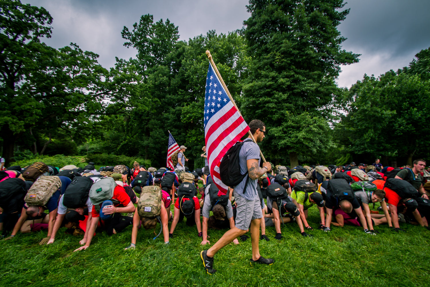 Man carrying an American flag walks past a group of people bending over on a grassy field in a park.