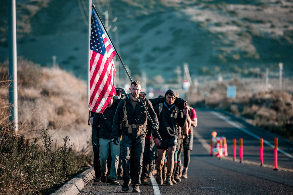 A group of people hike on a road; one person carries a large American flag at the front.