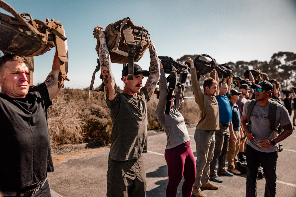 A group of people stand outdoors, lifting bags over their heads in a line on a sunny day.