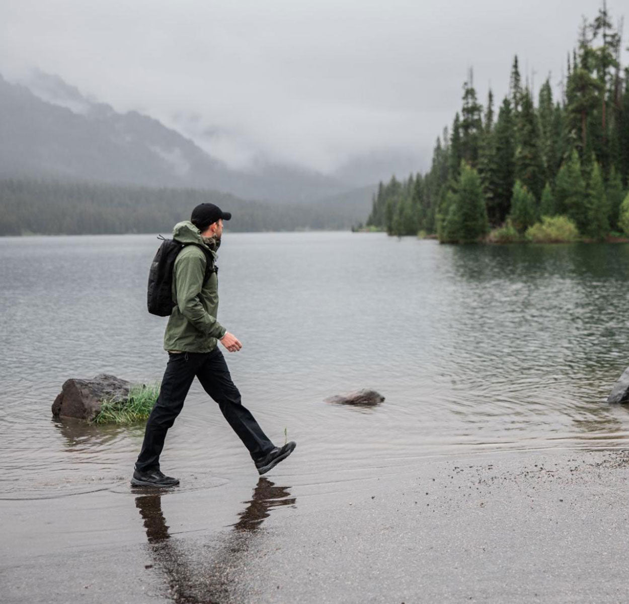 A person with a backpack walks by a misty lake surrounded by trees and mountains.