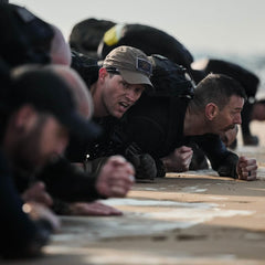 A group of men in military-style gear are performing a plank exercise on a sandy beach, their GORUCK Performance TAC Hats made with TOUGHDRY fabric handling the elements. They appear focused and determined, embodying teamwork and physical endurance in the challenging environment.