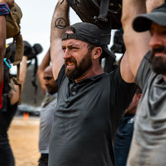 A group of people lift heavy bags overhead outside. The main focus is on a bearded man wearing a GORUCK Performance TAC Hat - Slick in TOUGHDRY material, along with a gray shirt. Other participants can be seen in the background, engaged in an outdoor fitness or team-building activity, highlighting resilience and teamwork.