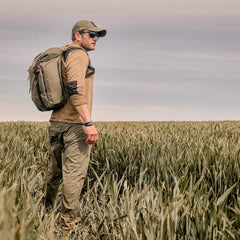 A man wearing a GORUCK Performance TAC Hat - TOUGHDRY, along with sunglasses and a backpack, stands in a field of tall grass under a cloudy sky, gazing into the distance. Dressed in outdoor gear made from ToughDry fabric, including a long-sleeved shirt and cargo pants, he's ready for his adventure.