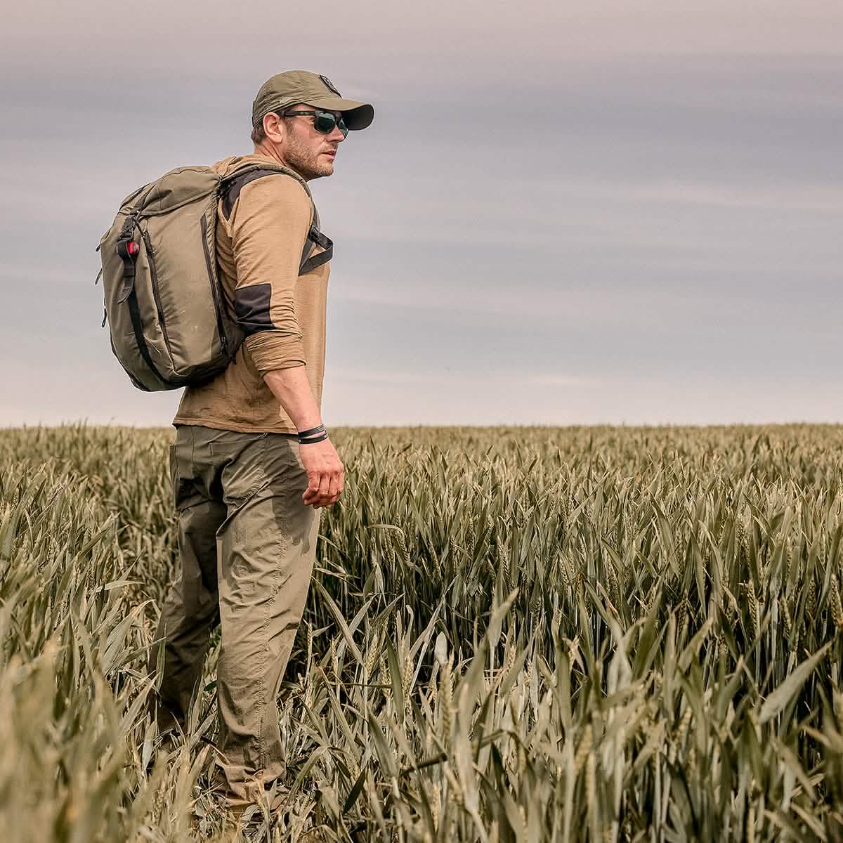 A man wearing a GORUCK Performance TAC Hat - TOUGHDRY, along with sunglasses and a backpack, stands in a field of tall grass under a cloudy sky, gazing into the distance. Dressed in outdoor gear made from ToughDry fabric, including a long-sleeved shirt and cargo pants, he's ready for his adventure.