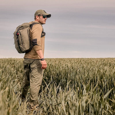A man wearing a GORUCK Performance TAC Hat - TOUGHDRY, along with sunglasses and a backpack, stands in a field of tall grass under a cloudy sky, gazing into the distance. Dressed in outdoor gear made from ToughDry fabric, including a long-sleeved shirt and cargo pants, he's ready for his adventure.