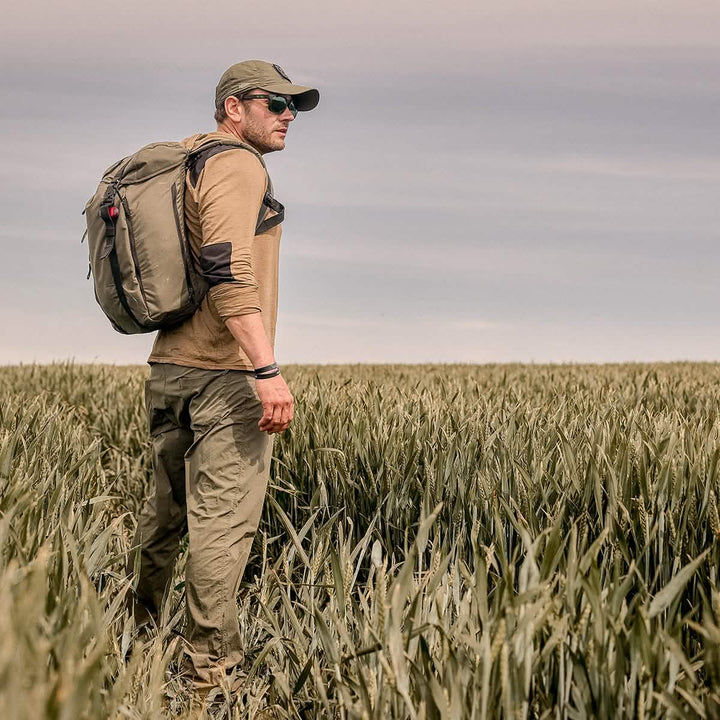 A man wearing a GORUCK Performance TAC Hat - TOUGHDRY, along with sunglasses and a backpack, stands in a field of tall grass under a cloudy sky, gazing into the distance. Dressed in outdoor gear made from ToughDry fabric, including a long-sleeved shirt and cargo pants, he's ready for his adventure.
