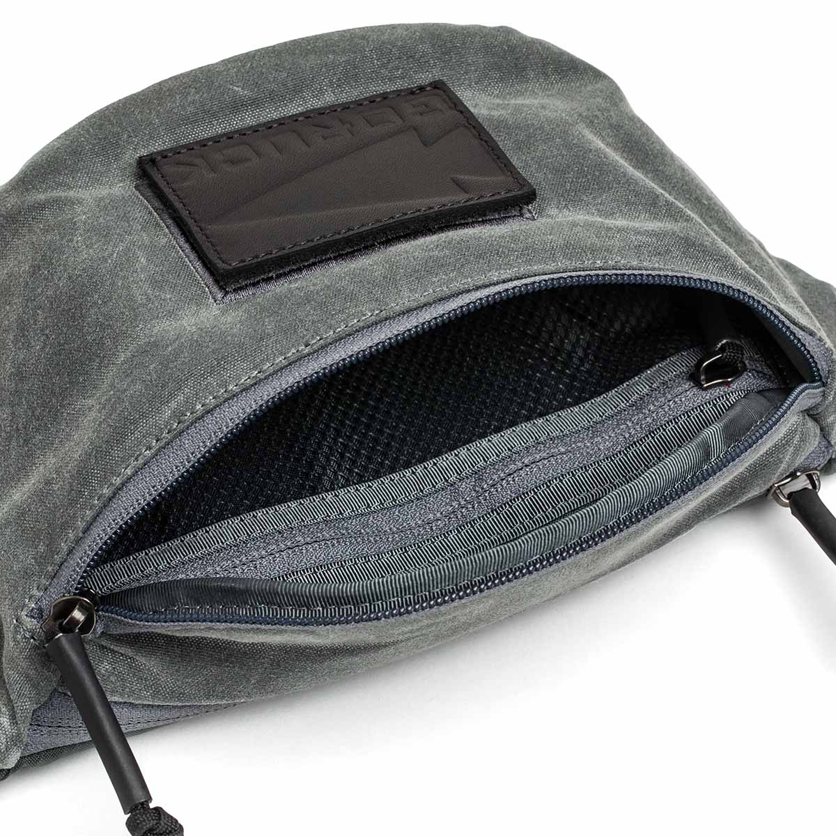 A close-up of the Tactical Fanny Pack - Waxed Canvas shows its open gray design with a black patch, silent zipper pulls, and visible black mesh lining inside.