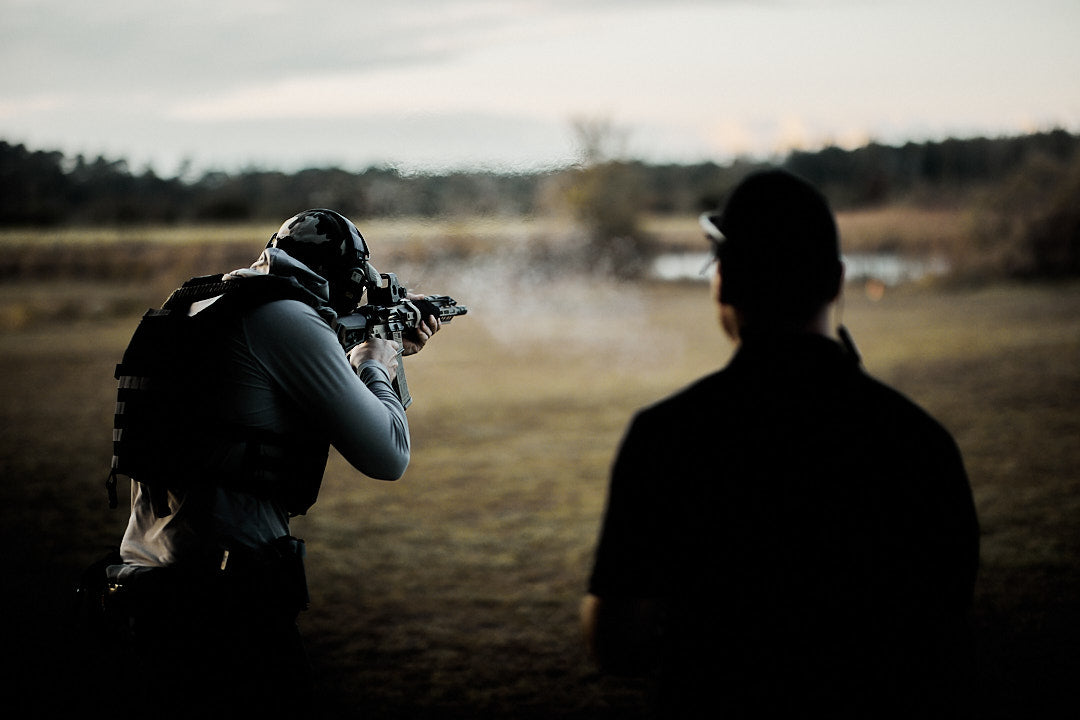 Person in tactical gear aiming a rifle outdoors, another person stands in the foreground, blurred.