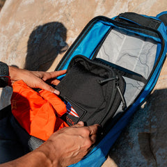 Hands packing a black GORUCK pouch and an orange item into a blue backpack with a topographic map interior
