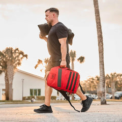Man walking outdoors carrying a red GORUCK rucksack and a black weighted sandbag at sunset