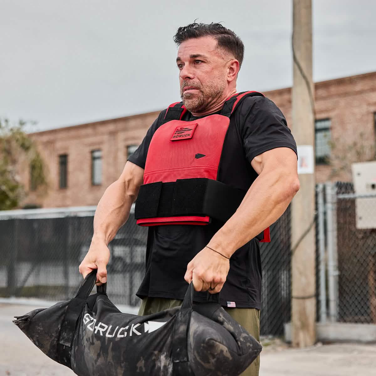 A man wearing the Men’s USA Performance Tee - ToughMesh lifts a heavy sandbag outdoors near a chain-link fence and brick building.