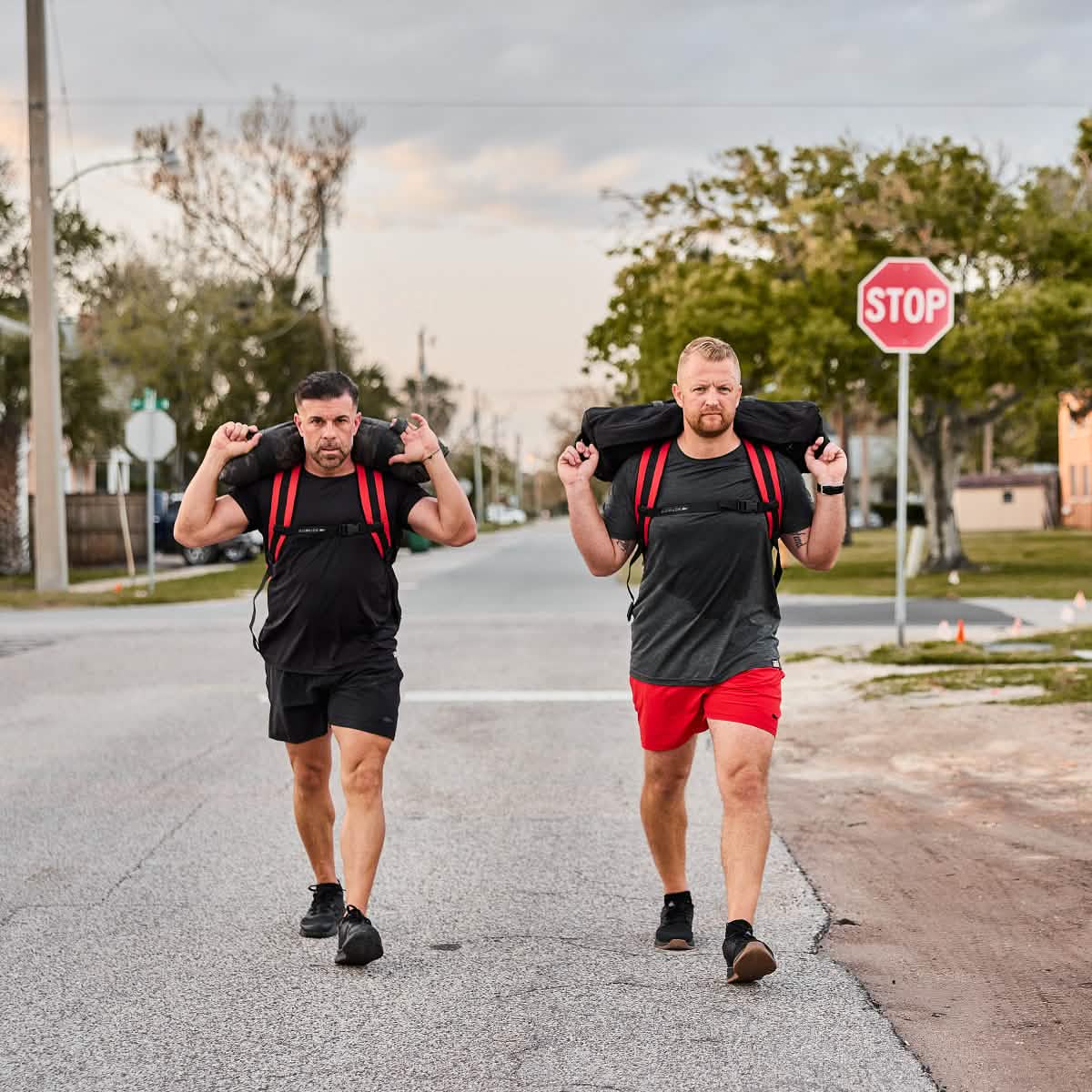 Two men walk down a street carrying sandbags on their shoulders, both wearing athletic clothes, including the Men’s USA Performance Tee - ToughMesh made from quick drying performance fabric.