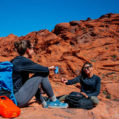 Two people sit on red rocks under a clear blue sky, clad in athletic gear and GORUCK Merino Challenge Socks - Trail Cuff. One carries a blue backpack, while the other holds a can; both are smiling as they enjoy their adventure in this desert-like paradise, confident in the Scars Lifetime Guarantee accompanying their journey.