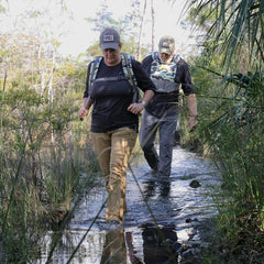 Two adventurers, clad in outdoor gear, navigate a shallow, muddy trail surrounded by lush foliage. The leader sports a backpack and Merino Challenge Socks - Trail Cuff from GORUCK for comfort, while the follower keeps pace. Their journey through nature speaks to the spirit of adventure and exploration.