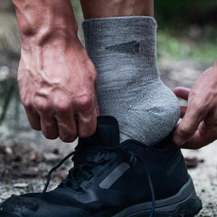 A person sporting GORUCK's Merino Challenge Socks - Trail Cuff in gray is adjusting their black hiking shoe, gearing up for a trek. The scene takes place outdoors on a dirt path, with an emphasis on the hands and shoes, capturing the adventurous spirit of the Trail Cuff and the confidence provided by Scars Lifetime Guarantee.