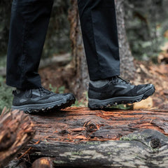 A person clad in black pants and Merino Challenge Socks - Trail Cuff by GORUCK strides across a fallen tree trunk in the forest. The socks, designed for rugged terrain, navigate the verdant surroundings with ease as trees and greenery envelop the serene scene.