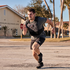 A man runs outdoors on a sunny day wearing the Training Weight Vest 2.0, athletic shorts, and sneakers.