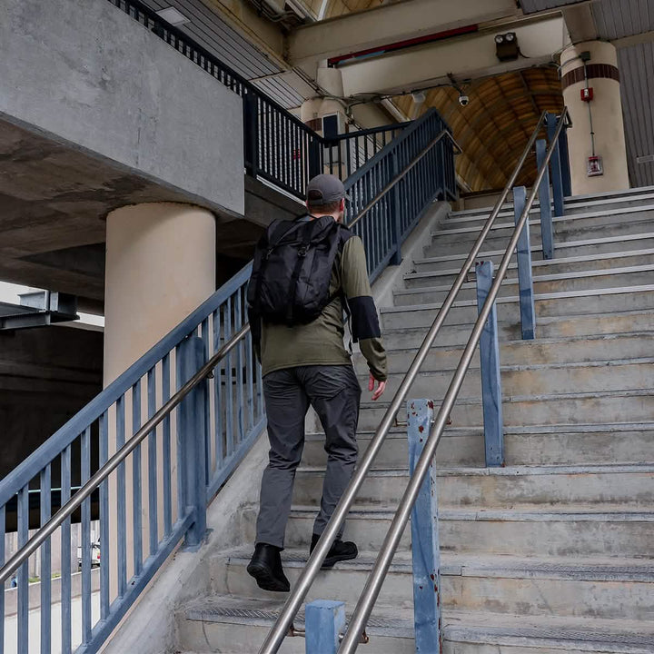 A person wearing a backpack, green jacket, gray pants, and a cap strides up a flight of concrete stairs in their MACV-1 Traveler footwear by GORUCK. The metal railings and visible beams under the sloped roof complement the tough, reliable grip of their All-Terrain Outsole.