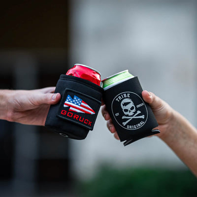 Two hands hold sleek black Beer Jackets, each displaying distinct logos. One showcases a GORUCK design with a U.S. flag, while the other features a skull and reads "Tribe Original." These Beer Jackets clink against softly blurred surroundings, adding flair to any gathering.