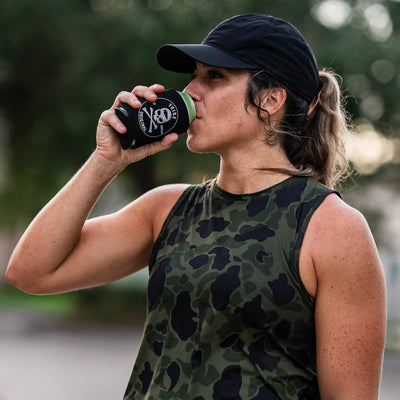 A woman wearing a camo tank top and black cap enjoys her drink from a black can featuring a skull and crossbones design, nestled in the GORUCK Beer Jacket - Tribe Original koozie. She stands outdoors with blurred greenery in the background.