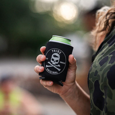 A person holds a canned beverage wrapped in a GORUCK Beer Jacket - Tribe Original, which features a skull and crossed bones design. The background is blurry, revealing hints of greenery and another person.