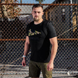Man in GORUCK Tropic Multicam Tee and rucksack standing by chain-link fence outdoors