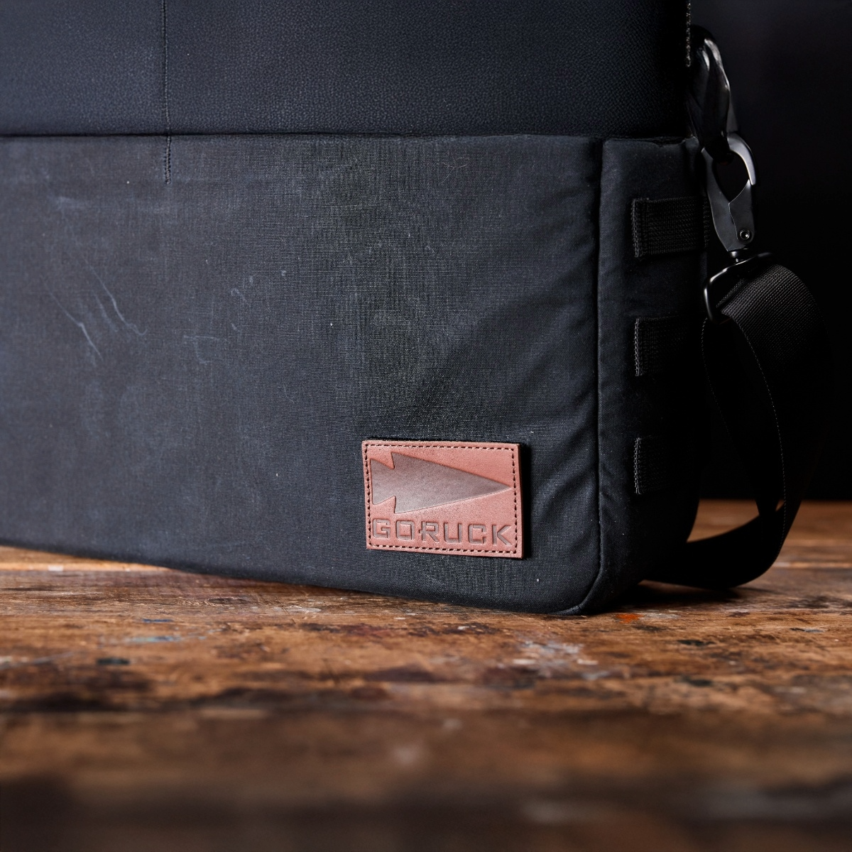 Close-up of a black Shoulder Bag - Waxed Canvas by GORUCK with a brown leather logo patch, designed for comfortable asymmetrical carries, shown on a wooden surface.