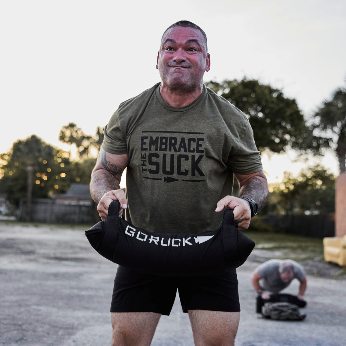 A man lifts a sandbag outdoors wearing the Embrace the Suck Tee - Tri-Blend, featuring the official USMC motto; another man is seen working out in the background.