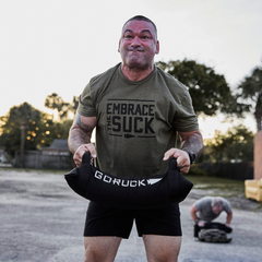 A man lifts a sandbag outdoors wearing the Embrace the Suck Tee - Tri-Blend, featuring the official USMC motto; another man is seen working out in the background.