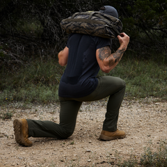 A man kneels outdoors with a camo-patterned sandbag on his shoulders, wearing boots and Men’s Simple Pants - Lightweight ToughDry® as part of his athletic gear.