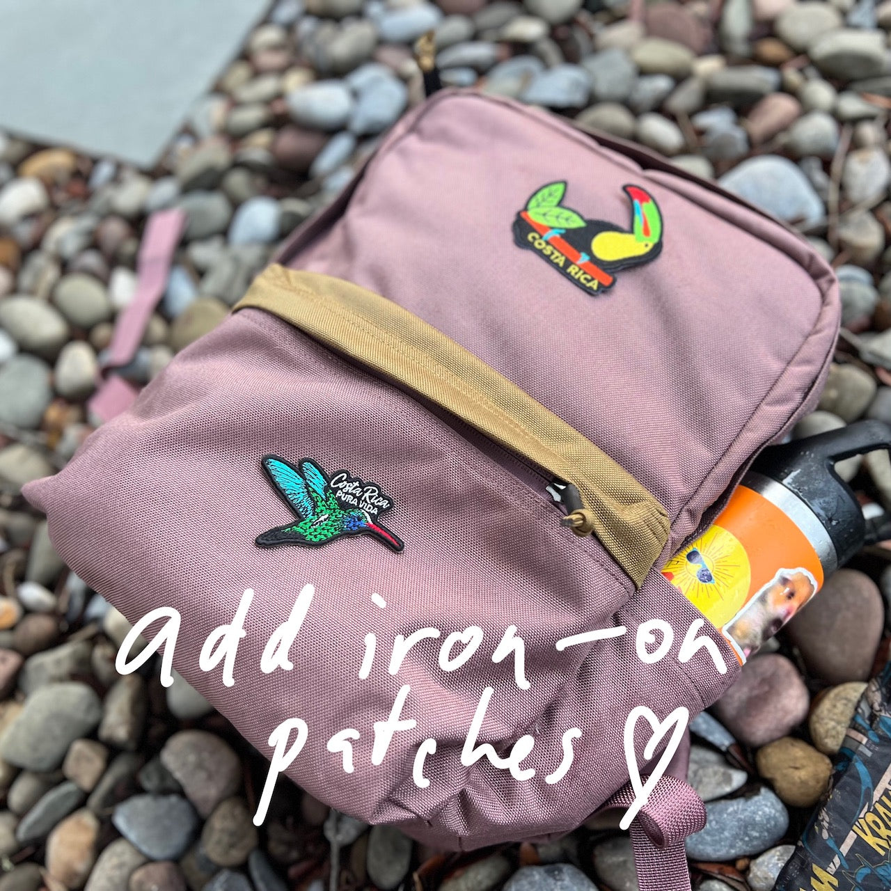 KR1 2.0 - Kid Ruck backpack in pink, featuring durable construction and colorful bird iron-on patches, with a water bottle in the side pocket, resting on pebbles. Text: add iron-on patches, make it yours.