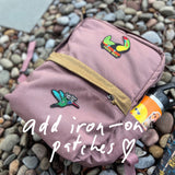 KR1 2.0 - Kid Ruck backpack in pink, featuring durable construction and colorful bird iron-on patches, with a water bottle in the side pocket, resting on pebbles. Text: add iron-on patches, make it yours.