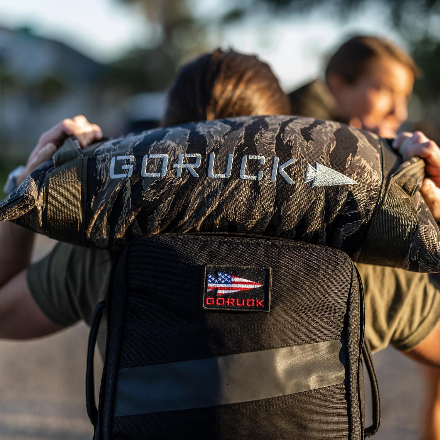 Person lifting a Simple Training Sandbag over their shoulders, GORUCK backpack with US flag patch visible—perfect for intense sandbag workouts in your home gym.