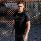 Man wearing GORUCK WWII Frogskin Camo tee and backpack standing by chain-link fence