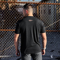 A man with short hair stands facing a chain-link fence, wearing the GORUCK Spearhead Tee - Tri-Blend in black. The T-shirt features a small spearhead logo on the back, and the sun casts intricate shadows from the fence onto the ground.