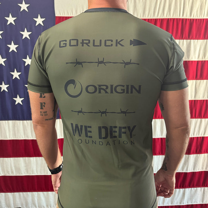 A man wearing the We Defy x GORUCK x Origin™ Rash Guard - Built By SCARS stands proudly in front of an American flag.