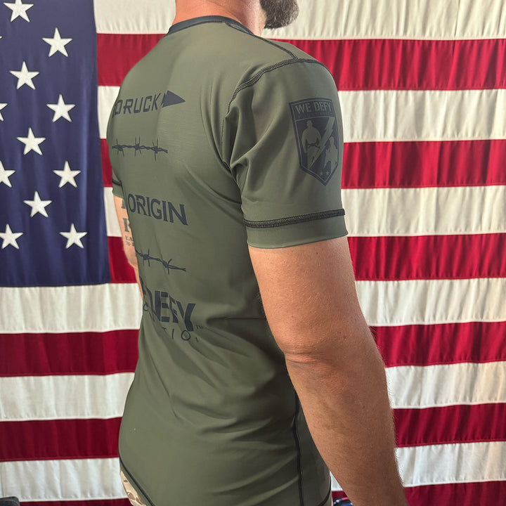 A man in a green shirt proudly stands before an American flag, showcasing the We Defy x GORUCK x Origin™ Rash Guard – Built By SCARS with its signature design on the back. This shirt is crafted with WARPWICK textile for ultimate comfort and UPF 50+ protection, seamlessly combining style and functionality.