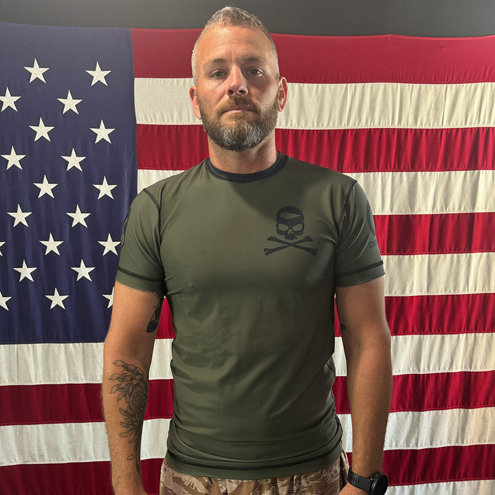 A man in a We Defy x GORUCK x Origin™ Rash Guard - Built By SCARS proudly stands in front of an American flag, enjoying the UPF 50+ skin protection it provides.