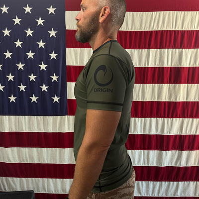 A man in a "We Defy x GORUCK x Origin™ Rash Guard - Built By SCARS," crafted from WARPWICK textile, stands sideways before an American flag backdrop.